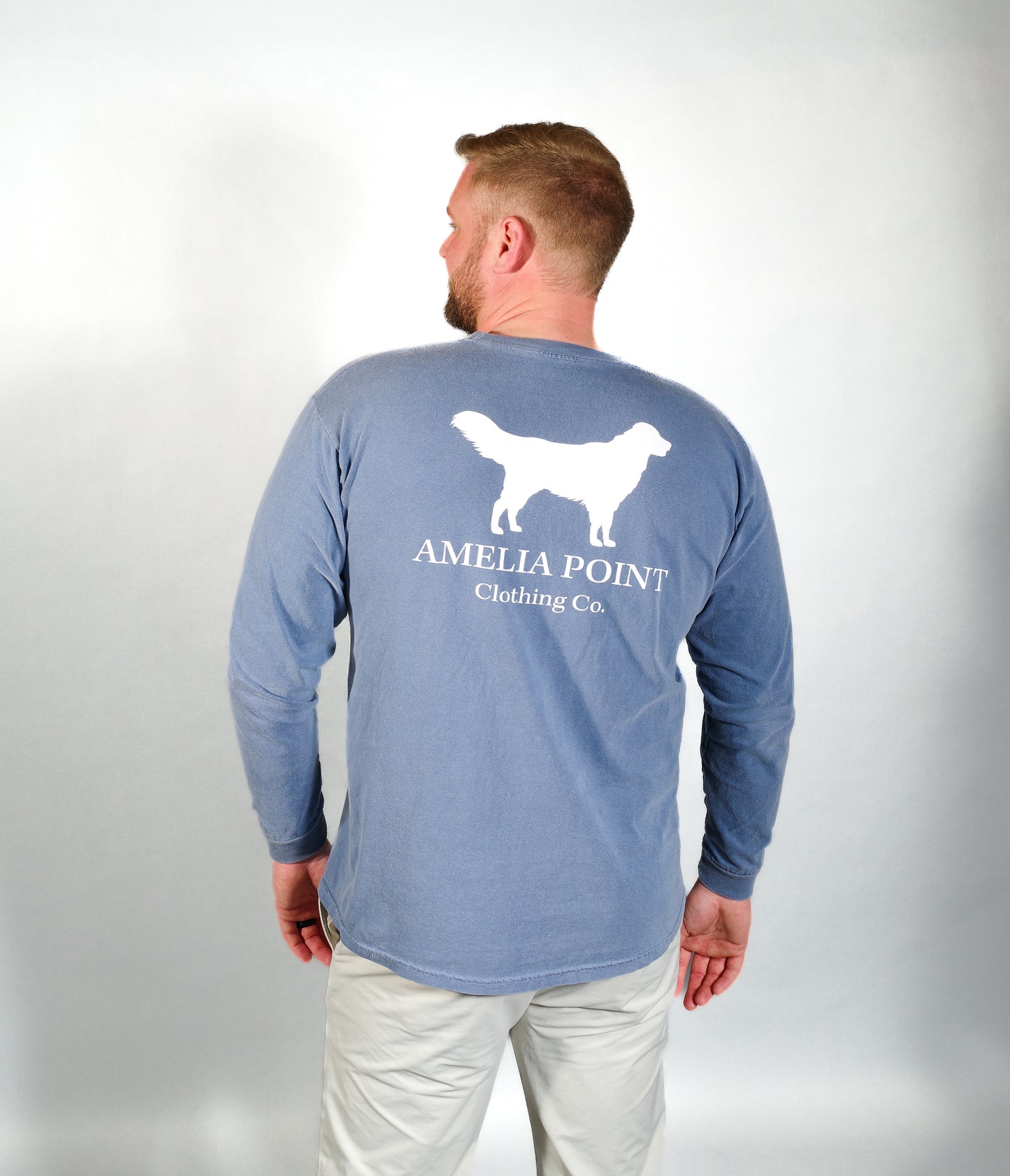 Up close image of the back side of the shirt on a male person, large golden retriever logo in white color on a blue jean color long sleeve shirt, Amelia Point Clothing Co is underneath the retriever logo