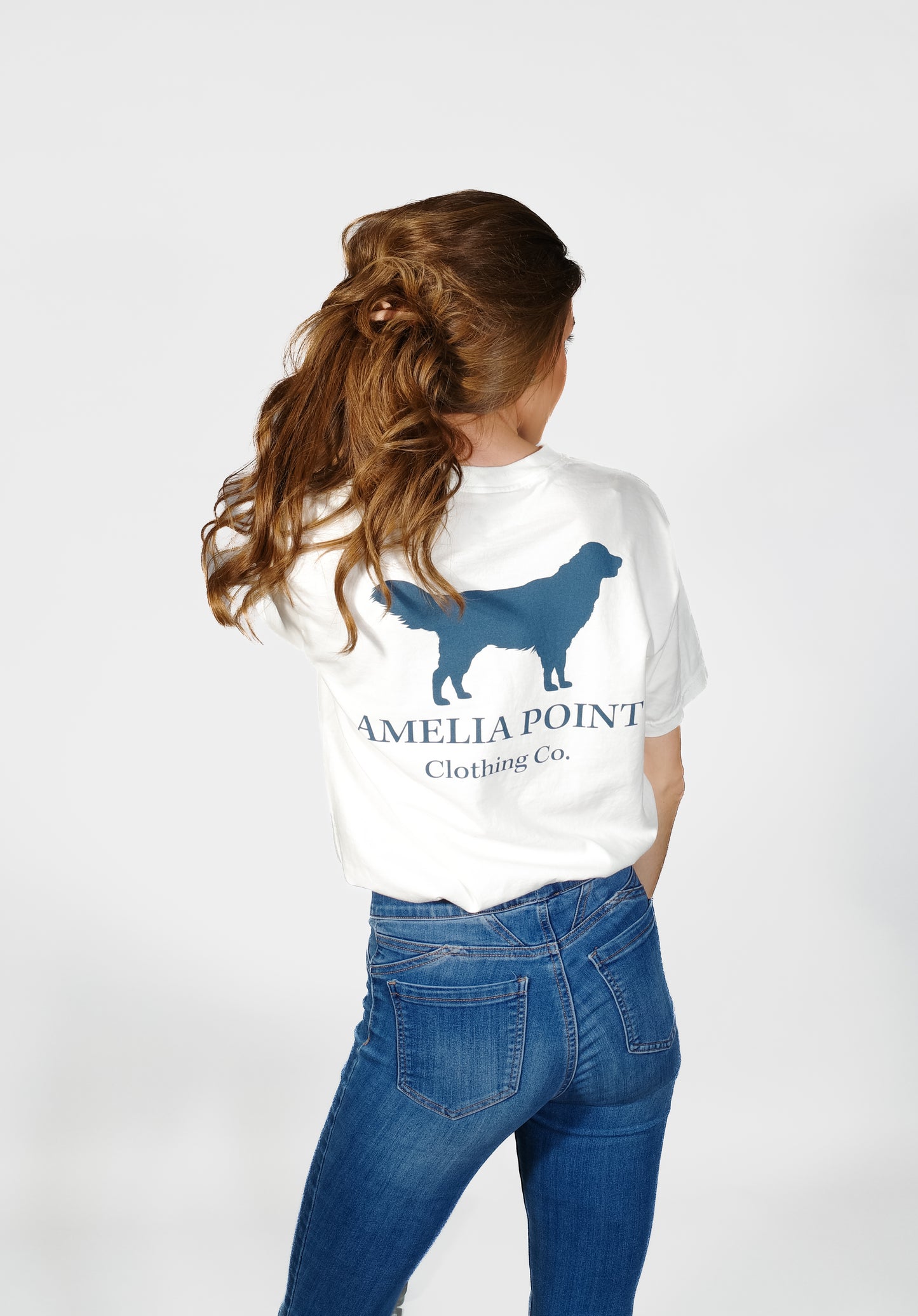 Up close image of the back side of the shirt on a female person, large golden retriever logo in true navy color on a white color short sleeve shirt, Amelia Point Clothing Co is underneath the retriever logo