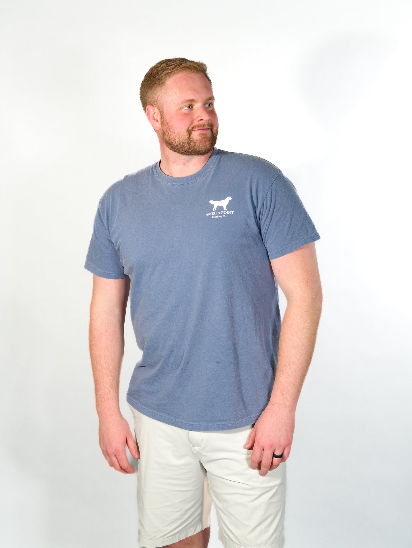 Image of the front side of the shirt on a male person, pocket sized golden retriever logo in white color on a blue jean color long sleeve shirt, Amelia Point Clothing Co is underneath the retriever logo