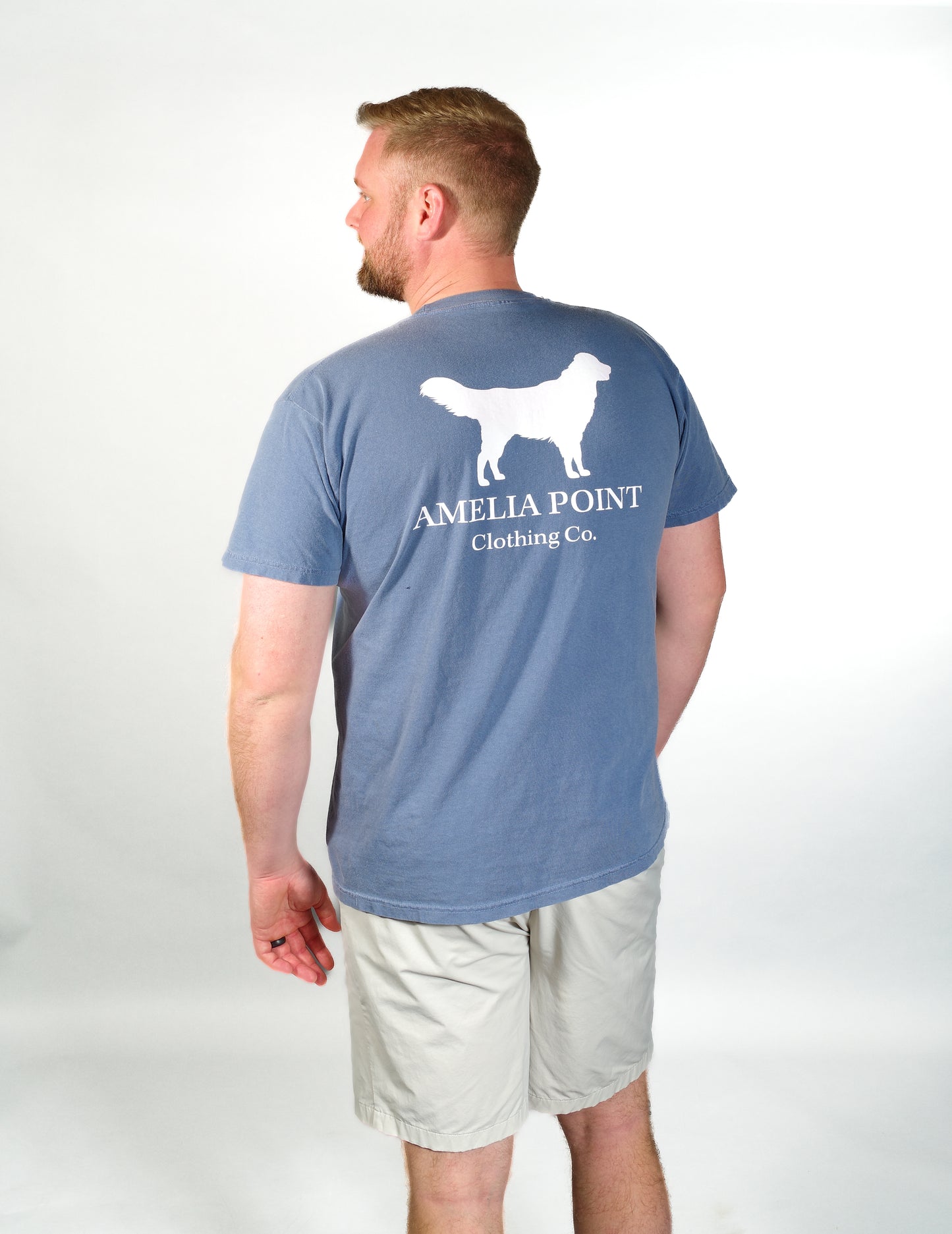 Up close image of the back side of the shirt on a male person, large golden retriever logo in white color on a blue jean color short sleeve shirt, Amelia Point Clothing Co is underneath the retriever logo