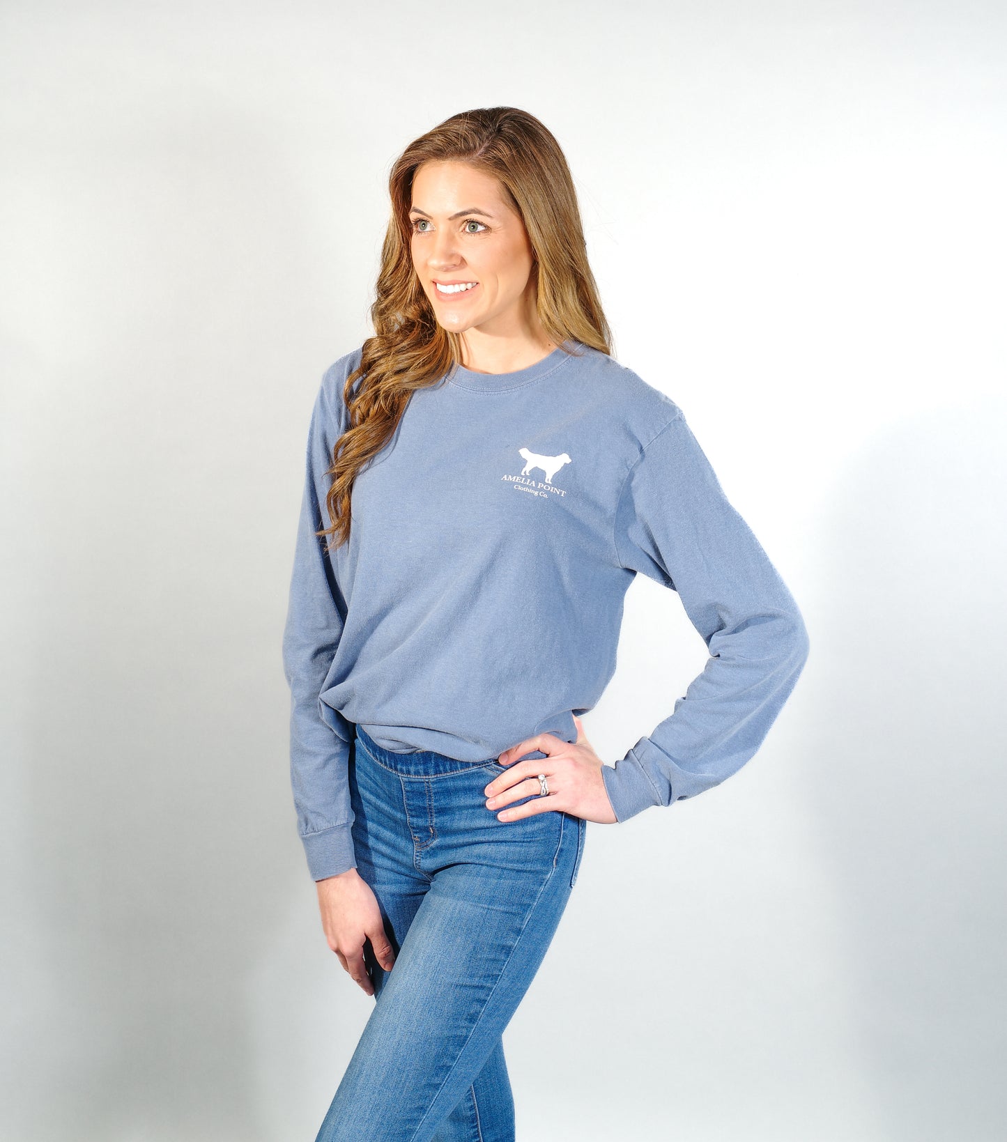 Image of the front side of the shirt on a female person, shirt pocket sized golden retriever logo in white color on a blue jean color long sleeve shirt, Amelia Point Clothing Co is underneath the retriever logo