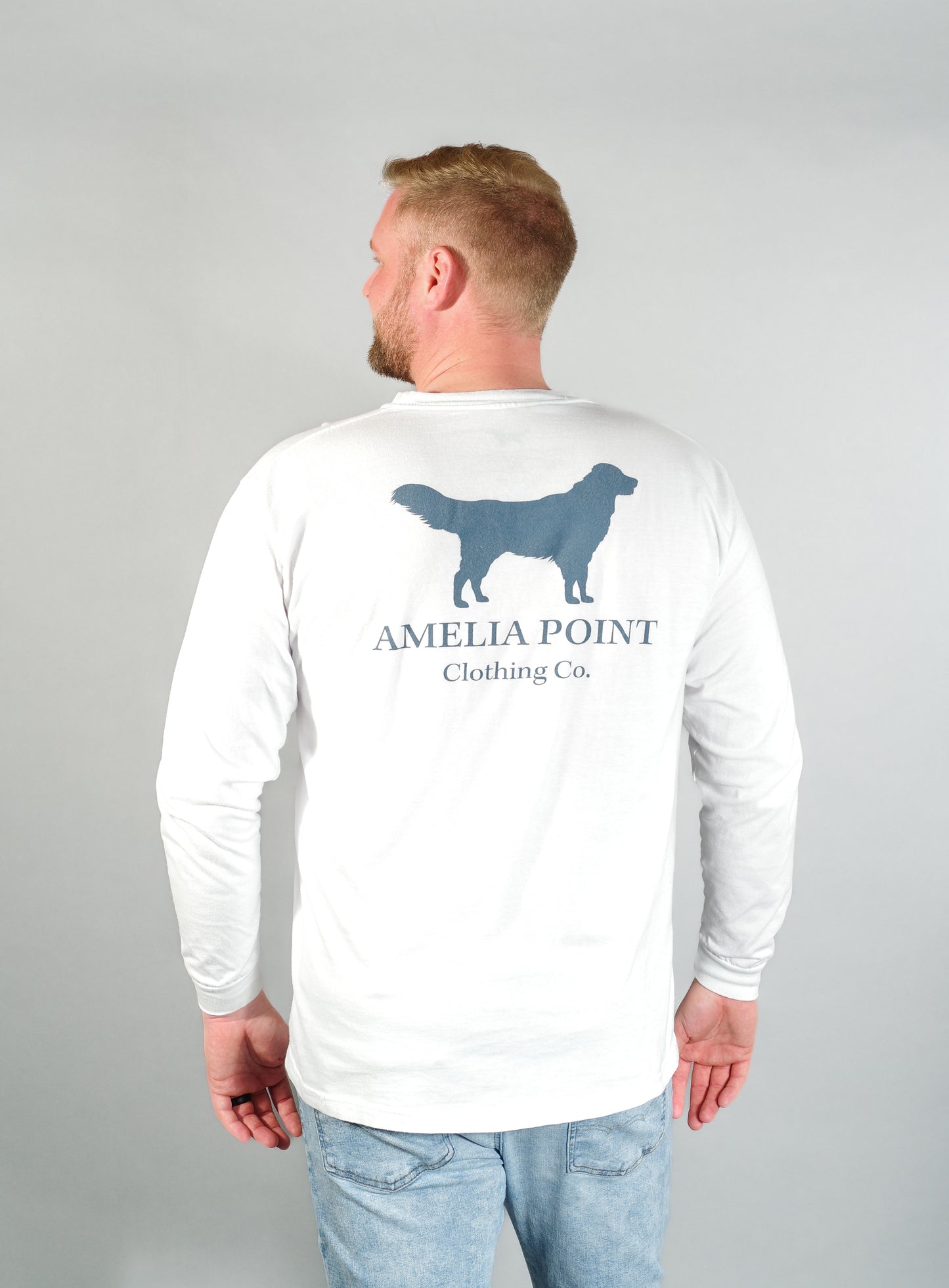 Up close image of the back side of the shirt on a male person, large golden retriever logo in true navy color on a white color long sleeve shirt, Amelia Point Clothing Co is underneath the retriever logo